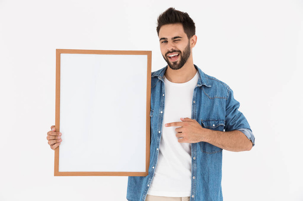 Image of smiling bearded man winking while holding and pointing finger at empty placard in frame for copyspace text - Foto, afbeelding