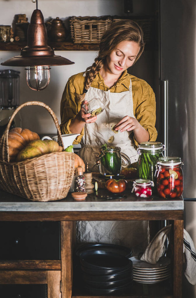 Autumn vegetable pickling and canning. Young blond woman in linen apron adding bay leaves to cucumbers and cooking homemade vegetables preserves in kitchen. Healthy organic fermented food concept - Foto, Imagem