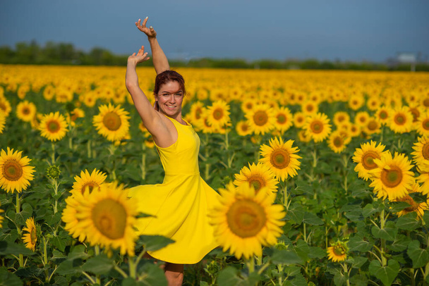 Red-haired woman in a yellow dress dancing with raised hands in a field of sunflowers. Beautiful girl in a skirt sun enjoys a cloudless day in the countryside. Pink locks of hair. - Foto, Imagem
