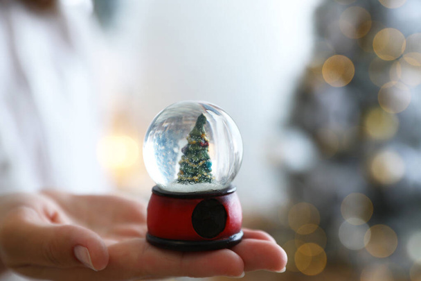 Young woman holding snow globe with Christmas tree on blurred background, closeup - Photo, Image