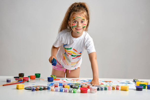Little girl in white t-shirt standing at table with whatman and colorful paints, painting on it with her hands. Isolated on white. Medium close-up. - Photo, Image