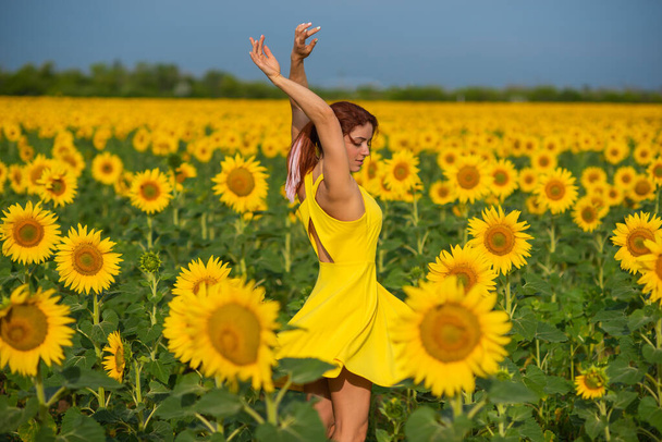 A red-haired woman in a yellow dress is standing in a field of sunflowers. Beautiful girl in a skirt sun enjoys a cloudless day in the countryside. Pink locks of hair. - Foto, afbeelding