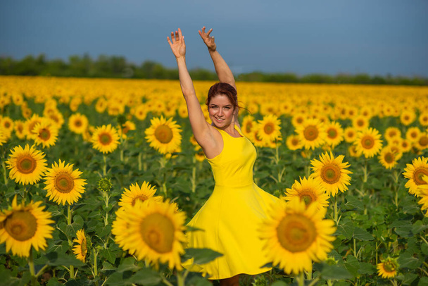 Red-haired woman in a yellow dress dancing with raised hands in a field of sunflowers. Beautiful girl in a skirt sun enjoys a cloudless day in the countryside. Pink locks of hair. - Fotó, kép