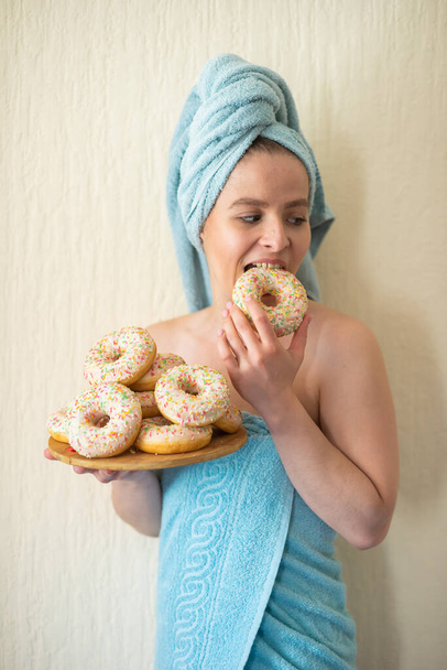 The girl in a towel has breakfast in the bed. A young attractive woman eats donuts in bed. The girl with a towel on her head happily eats donuts. - Foto, imagen