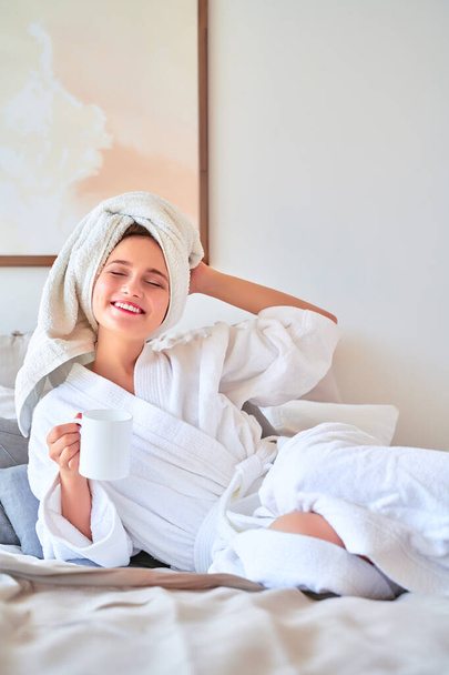 Photo of happy woman in white bathrobe with mug of tea in her hands lying on bed. - 写真・画像