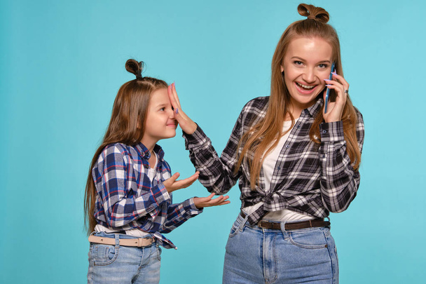 Mom and daughter dressed in checkered shirts and blue denim jeans are using smartphone while posing against a blue studio background. Close-up shot. - 写真・画像