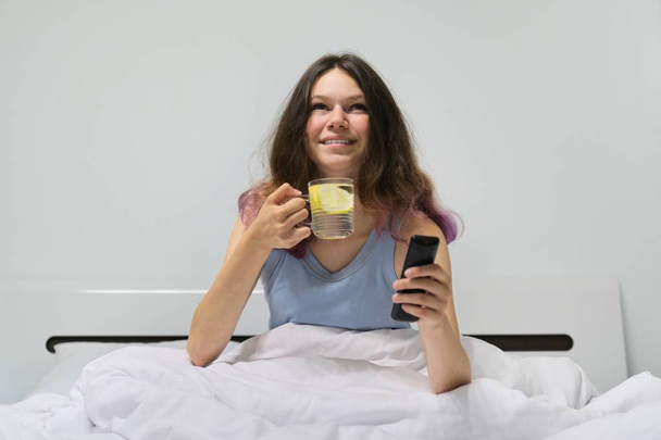 Teen girl sitting in bed with glass of water with lemon holding TV remote in hand - Photo, Image