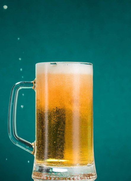 Glass of beer with foam and spray from falling ice on a turquoise green background - Foto, afbeelding
