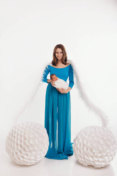 A girl in a blue dress with angel wings holding a newborn baby on a white background. An angel-woman and child have big white balls. The concept of Christmas, mystical creatures, Halloween - Foto, imagen