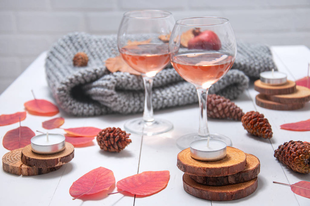 小さなキャンドル、バラのワインと2つのメガネ、コーン、乾燥した赤い葉、白い木製のテーブルの上に編んだ灰色のスカーフ。こんにちは、秋。秋の風物詩. - 写真・画像