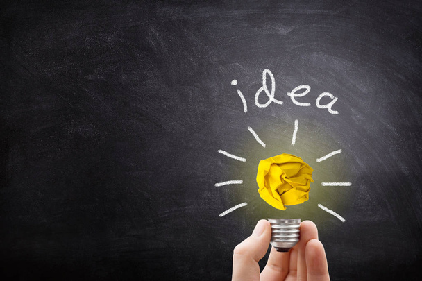 Business Idea Concept : Hand holding yellow crumpled paper ball light bulb lighting grow around on chalkboard. - Photo, Image