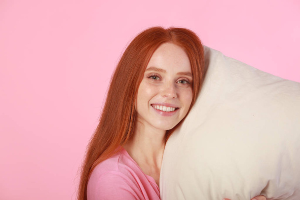 beautiful young woman with red hair with a pillow in hands on a pink background - Fotografie, Obrázek