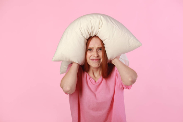 beautiful young woman with red hair with a pillow on her head on a pink background - 写真・画像