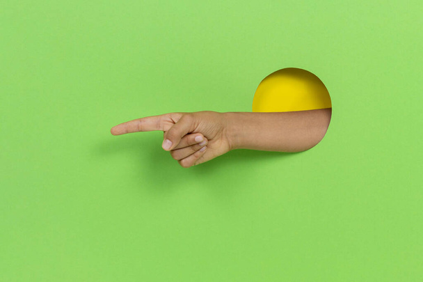 Hand door gat in licht groene achtergrond wijzend met de vinger naar iets, het tonen van richting teken - Foto, afbeelding