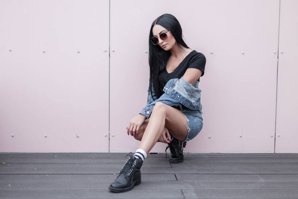 Pretty fashionable young in sunglasses in a trendy blue skirt in a stylish black top in a denim jacket in leather boots relaxes near a wall on an outdoor terrace. American modern urban hipster girl. - Fotografie, Obrázek