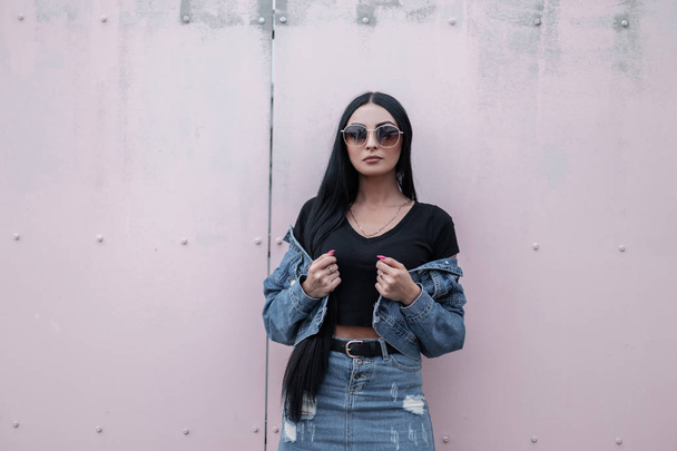 Mulher hipster bonita moderna em óculos de sol pretos elegantes fica e endireita uma jaqueta de moda jeans perto de uma parede vintage. Modelo de menina morena atraente em roupas da moda posando ao ar livre
. - Foto, Imagem