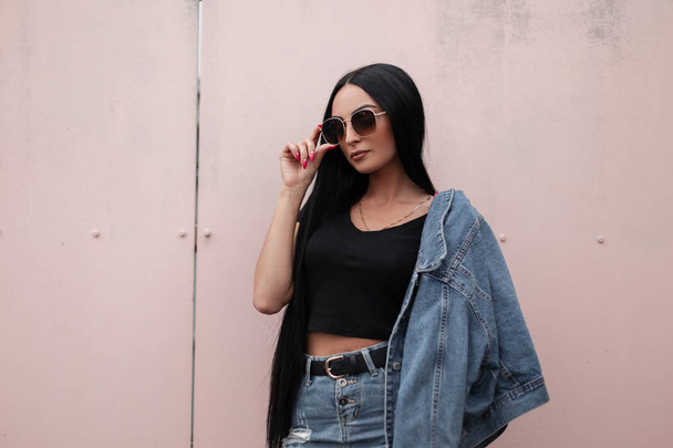 attraktive junge brünette Frau mit einem trendigen schwarzen Oberteil in einer stylischen Jeansjacke in einem blauen Jeansrock im Freien in der Nähe des Gebäudes posiert. schönes Hipster-Mädchen richtet Sonnenbrille auf. - Foto, Bild