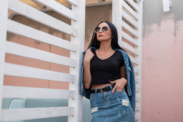 Beautiful sexy young hipster woman in a stylish denim jacket in a trendy black top in a vintage jeans skirt in sunglasses resting near a wooden wall in the city. Urban brunette girl with long hair. - Photo, Image