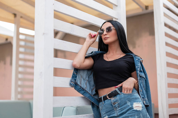 Die modische amerikanische junge Hipsterfrau in einem stylischen schwarzen Top in einer Vintage-Jeansjacke in einem trendigen Rock ruht sich in der Stadt an einer weißen Holzwand aus. attraktives Mädchen-Modell. - Foto, Bild