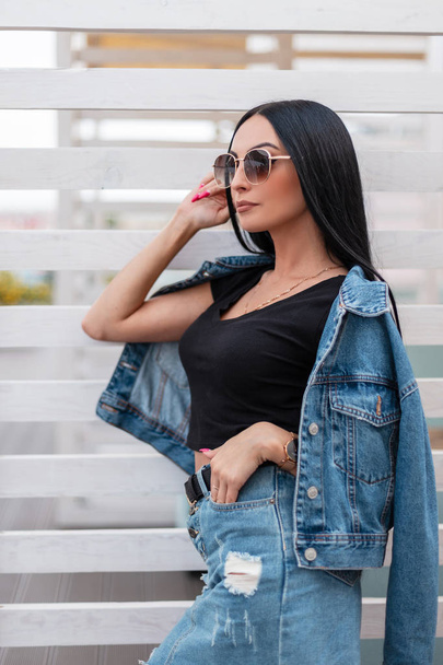 Pretty urban young woman fashion model with luxurious black hair in trendy blue denim clothes in stylish dark sunglasses posing outdoors near a white wooden wall. Beautiful hipster girl in the city. - Photo, Image