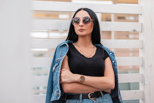 Pretty young brunette woman with black hair in a fashionable denim jacket in a vintage skirt in a stylish top in sunglasses posing near a wooden wall outdoors on a summer day. American hipster girl. - Foto, imagen