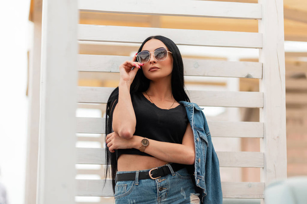 Stylish young woman model with black long hair in fashionable jeans clothes straightens trendy sunglasses. Attractive girl hipster posing near a vintage wooden white wall outdoors. - Photo, Image