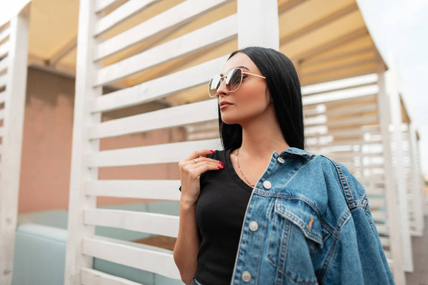 Model of a young American woman with black hair in a trendy black t-shirt in fashionable jeans jacket in a stylish sunglasses. Urban girl hipster stands near a vintage wooden white wall outdoors. - Photo, Image