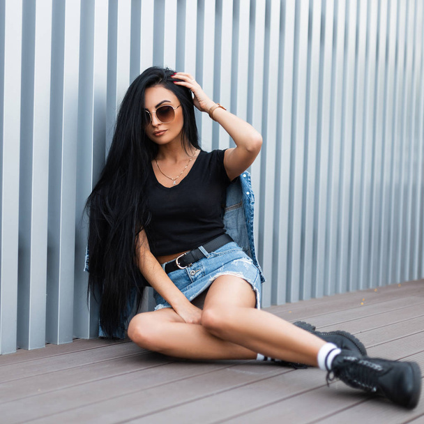 Beautiful young hipster woman in a trendy denim jacket in a blue skirt in a T-shirt in stylish leather black boots in sunglasses sitting on the floor near the metal wall.Urban girl with chic long hair - Photo, Image