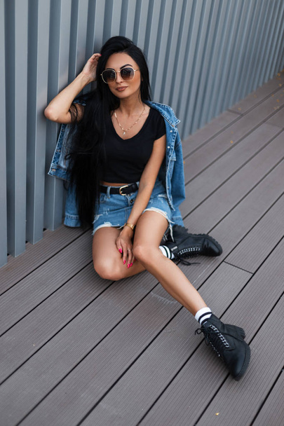 Trendy hippe jonge vrouw met lang haar in modieuze jeans kleding in stijlvol zwart lederen laarzen zit in de stad vlakbij een zilveren metalen muur. Amerikaans meisje rust buiten. Stijlvolle dameskleding. - Foto, afbeelding