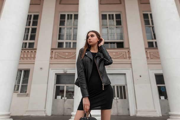Die hübsche junge Frau in einer schicken schwarzen Lederjacke im eleganten Kleid mit stylischer Tasche steht neben einer weißen Säule auf der Straße. modernes europäisches Mädchen. Freizeitmode im Herbst. - Foto, Bild