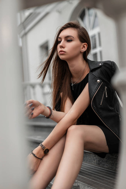 Modelo europeo de mujer joven en un elegante vestido negro corto en una elegante chaqueta de cuero negro se encuentra en una escalera de piedra cerca de un moderno edificio blanco. Hermosa chica descansa en la ciudad en un día de otoño
. - Foto, Imagen