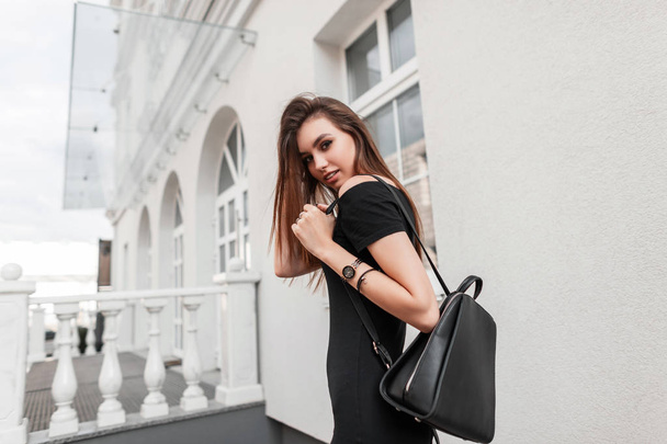 Piękny model młodej kobiety w eleganckiej czarnej sukience ze skórzanym stylowym czarnym plecakiem podróżuje po mieście. Cute modne dziewczyna cieszy się spacerem po ulicy. Uliczny styl młodzieży. - Zdjęcie, obraz