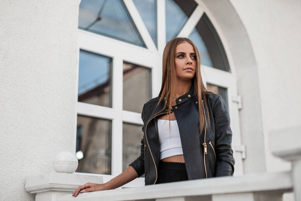 Fashionable young woman with long brown hair in a stylish black leather jacket in a white vintage blouse stands near a vintage white wall in the city. Pretty modern girl fashion model rests outdoors - Photo, Image