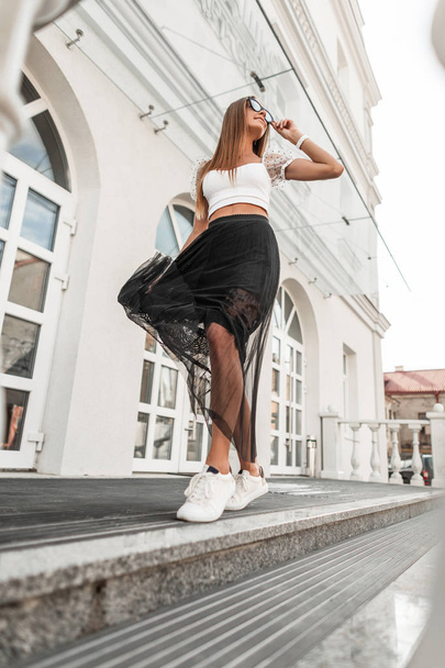 Mooie jonge vrouw met glimlach in een stijlvolle zwarte rok in trendy zonnebril in een top in modieuze schoenen poseren buiten in de buurt van een modern gebouw buiten. Aantrekkelijk gelukkig hipster meisje in de straat. - Foto, afbeelding