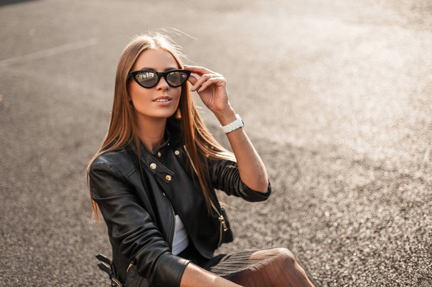 Cheerful pretty young hipster woman in a stylish leather jacket in an elegant black skirt is resting on the asphalt on a sunny autumn day. Happy girl with a cute smile straightens trendy sunglasses. - Fotoğraf, Görsel