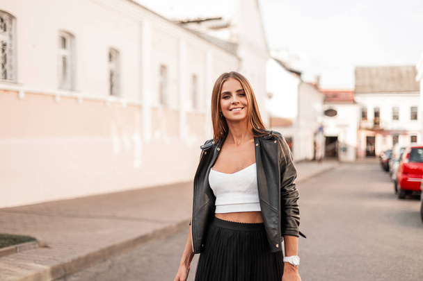 Pretty positive young woman in a stylish black leather jacket in fashionable white shirt walks on the street near vintage buildings. Trendy happy girl model relaxes outdoors. Youth womens fashion. - Photo, Image