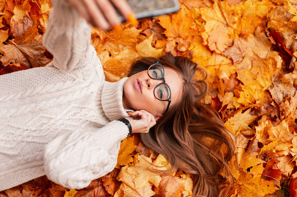 ファッショナブルなニットセーターのスタイリッシュな眼鏡で魅力的なかなり若いヒップスター女性は公園のオレンジの葉にあり、電話で自撮りをします。美しい少女は休みです。上からの眺め. - 写真・画像