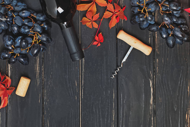 Uvas, vino tinto en botella y hojas de otoño sobre la mesa de madera negra
 - Foto, Imagen