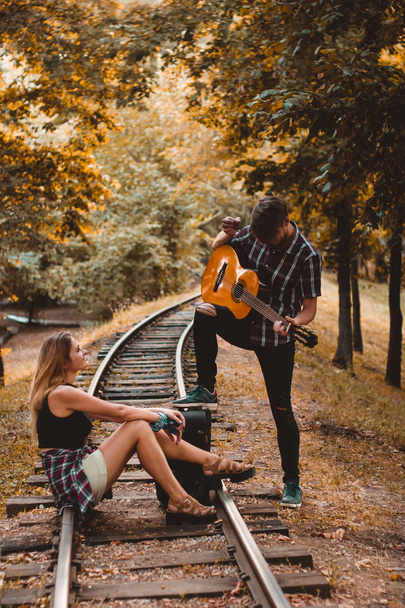 Δυο νεαροί εραστές έχασαν το τρένο. Παίζοντας τραγούδι με κιθάρα στις ράγες στο δάσος του φθινοπώρου περιμένοντας το επόμενο τρένο. - Φωτογραφία, εικόνα