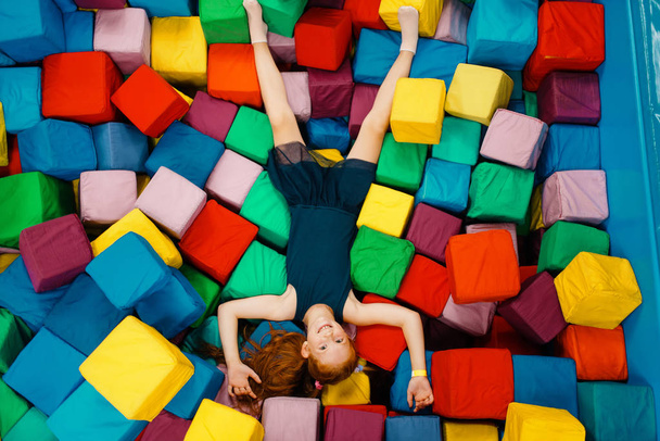 Мила дівчинка лежить у м'яких кубиках, дитячий майданчик у розважальному центрі. Ігрова зона в приміщенні, ігрова кімната
 - Фото, зображення