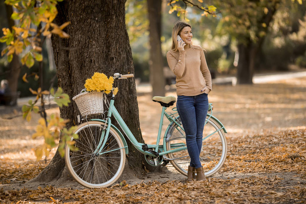 秋の公園でスマートフォンを使って自転車を使う可愛い若い女性 - 写真・画像
