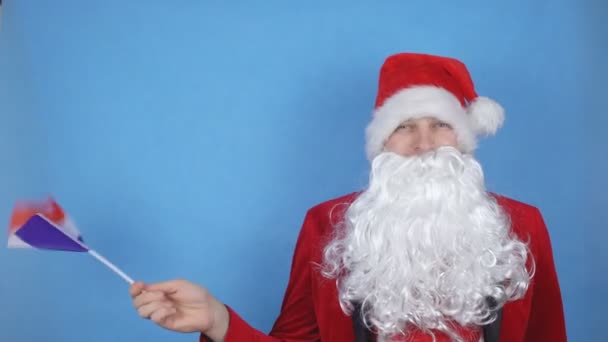 A man in a Santa Claus costume with a beard waving the flag of France on a blue background. New Year holidays concept in the world - Video, Çekim