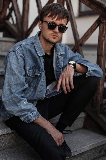 Ein junger Mann in blauer Jeansjacke, schicker Hose und stylischer schwarzer Sonnenbrille sitzt an einem Frühlingstag auf einer Vintage-Treppe auf der Straße. modischer Typ Hipster genießt Erholung im Freien - Foto, Bild