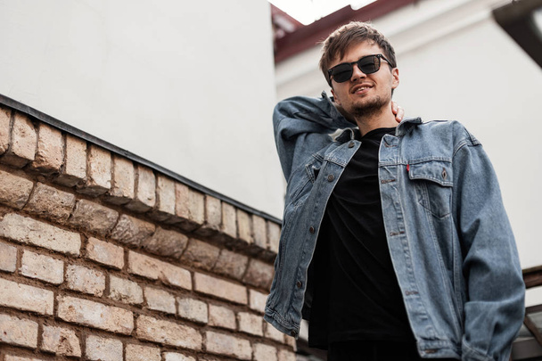 Cute American young man hipster in a vintage blue denim jacket in a black T-shirt in stylish sunglasses posing near a vintage building in the city. Attractive positive guy with a cute smile outdoors. - Fotoğraf, Görsel