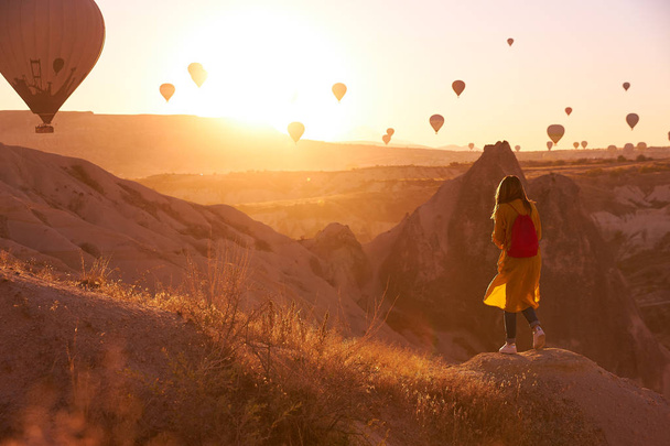 sunrise photo in Cappadocia with air balloons in the sky and a girl with red bag dark jeans yellow cardican and white sneakers standing on the hill and watching the view - Valokuva, kuva