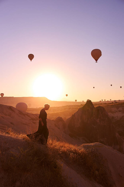 zdjęcie wschodu słońca w Kapadocji z balonami na niebie i dziewczyną chodzącą po wzgórzu i obserwującą ziemię - Zdjęcie, obraz