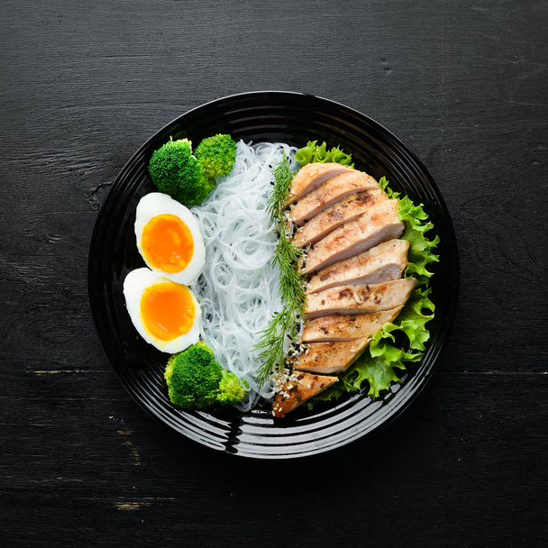Rice noodles with chicken fillet, egg and broccoli in a black plate on a white wooden background. Top view. Free space for your text. - Zdjęcie, obraz