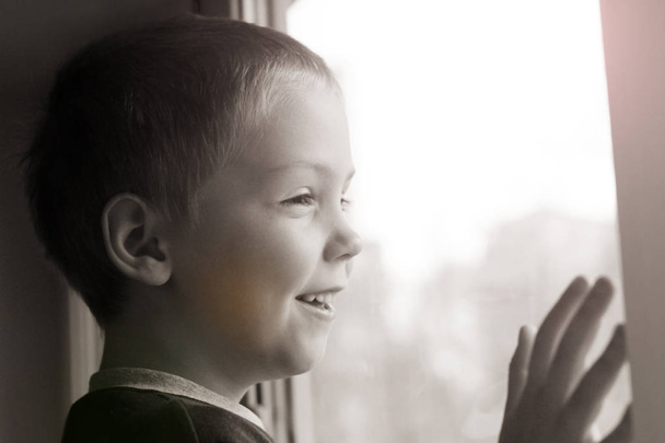 Το ξανθό αγόρι χαμογελάει και κοιτάζει έξω από το παράθυρο. - Φωτογραφία, εικόνα