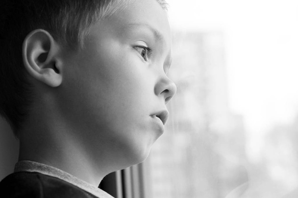 Το ξανθό αγόρι είναι λυπημένο και κοιτάζει έξω από το παράθυρο. - Φωτογραφία, εικόνα