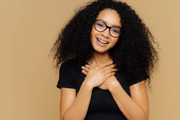 Touched lovely woman with curly hairstyle, keeps palms on chest, tilts head, has charming smile on face, wears optical glasses, black casual t shirt, models against brown background. Gratitude - Fotografie, Obrázek